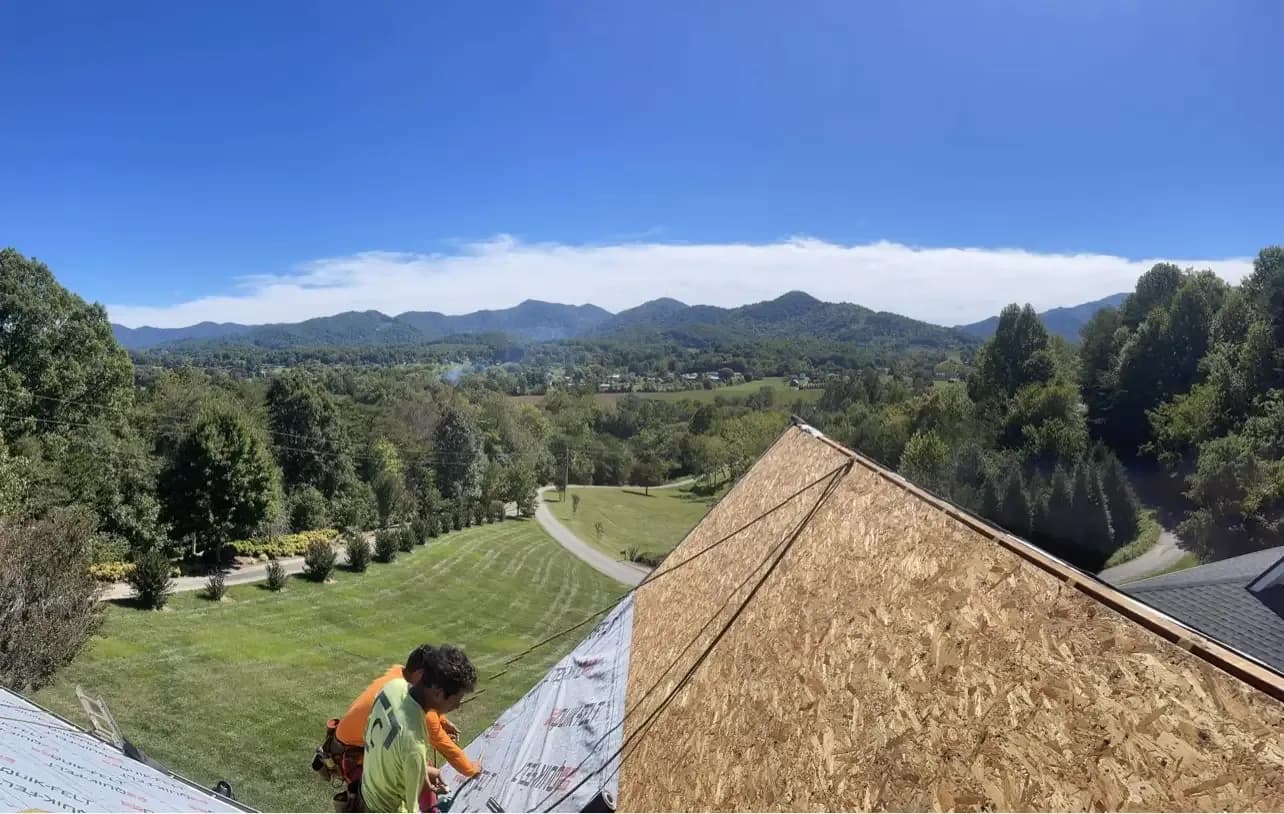 Roofers work on repairing a roof. Green hills are visible in the distance.