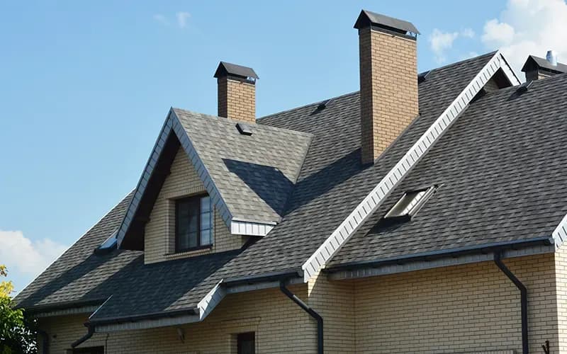 An asphalt shingle roof on a light brown brick house with dark gutters and downspouts and two chimneys