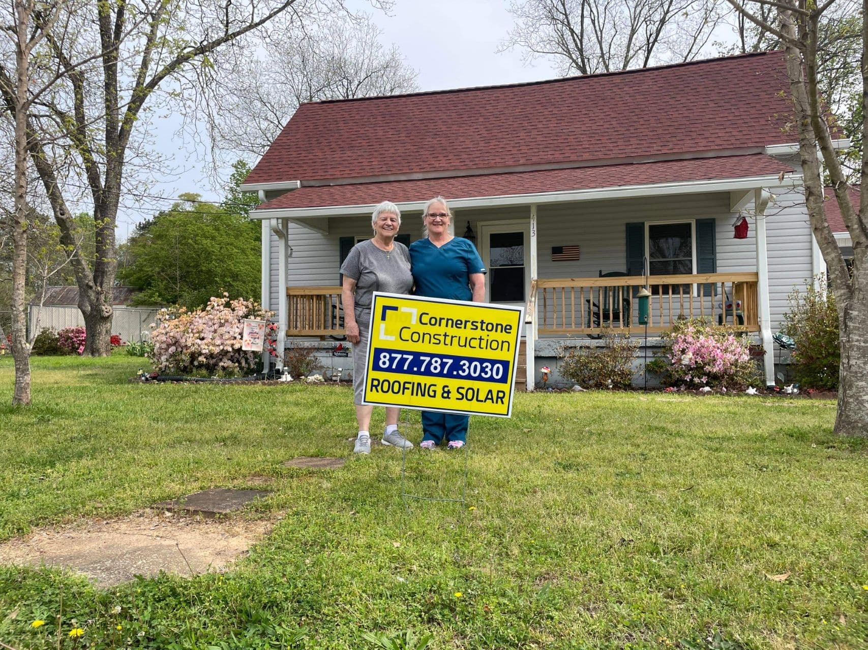 Homeowners stand behind a Cornerstone Construction lawn sign in front of a light gray house with new shingle roofing.